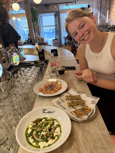 A cheerful person smiles at the camera while enjoying a variety of dishes and drinks at a modern bar